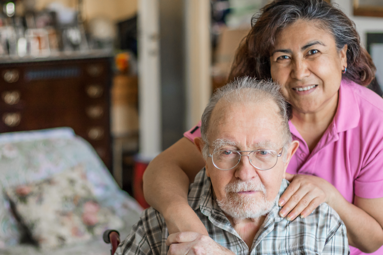 a woman standing behind an older man with one hand on his shoulder and the other arm hanging down holding the man's hand