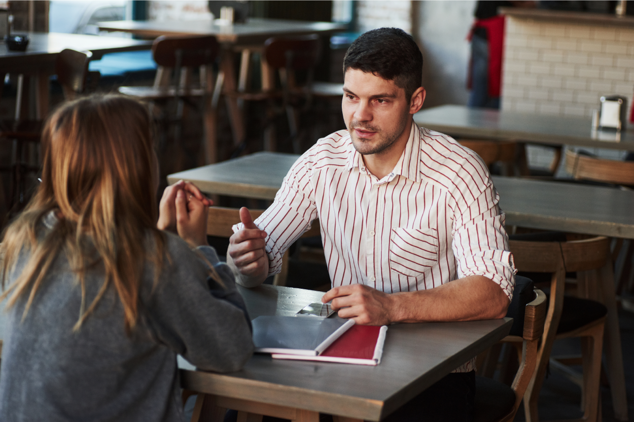 A person with their back to the camera sits at a table with a man. They are engaged in a conversation. Two notebooks are on the table in front of them.