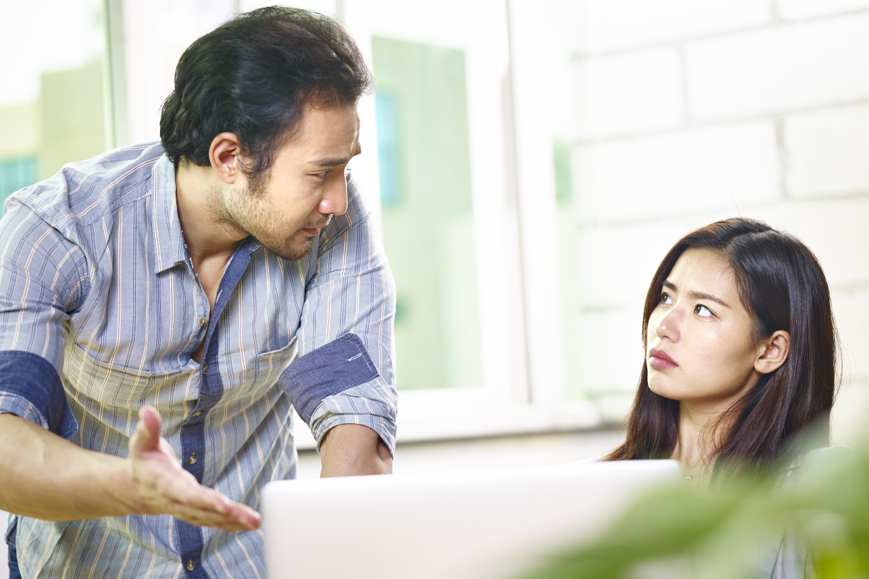 Man standing over a woman at a work desk reprimanding her.