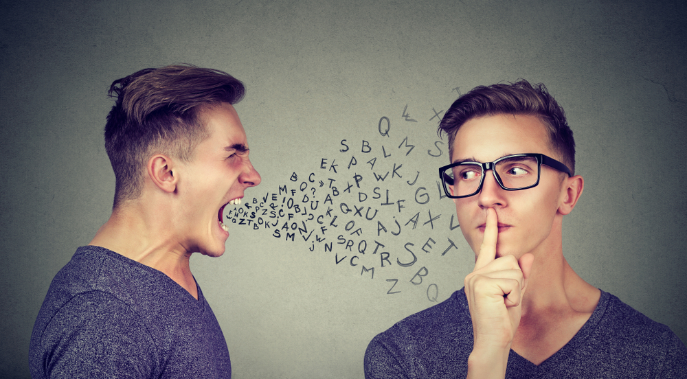 Man shouting with abusive words at another man who has one finger on his lips asking for silence