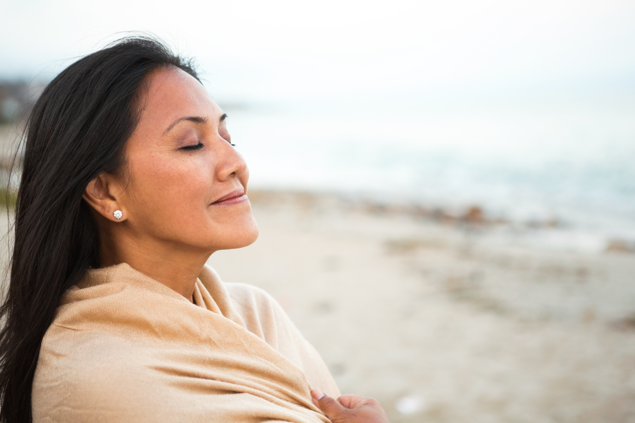 A woman facing the ocean with her eye closed and taking a deep breath