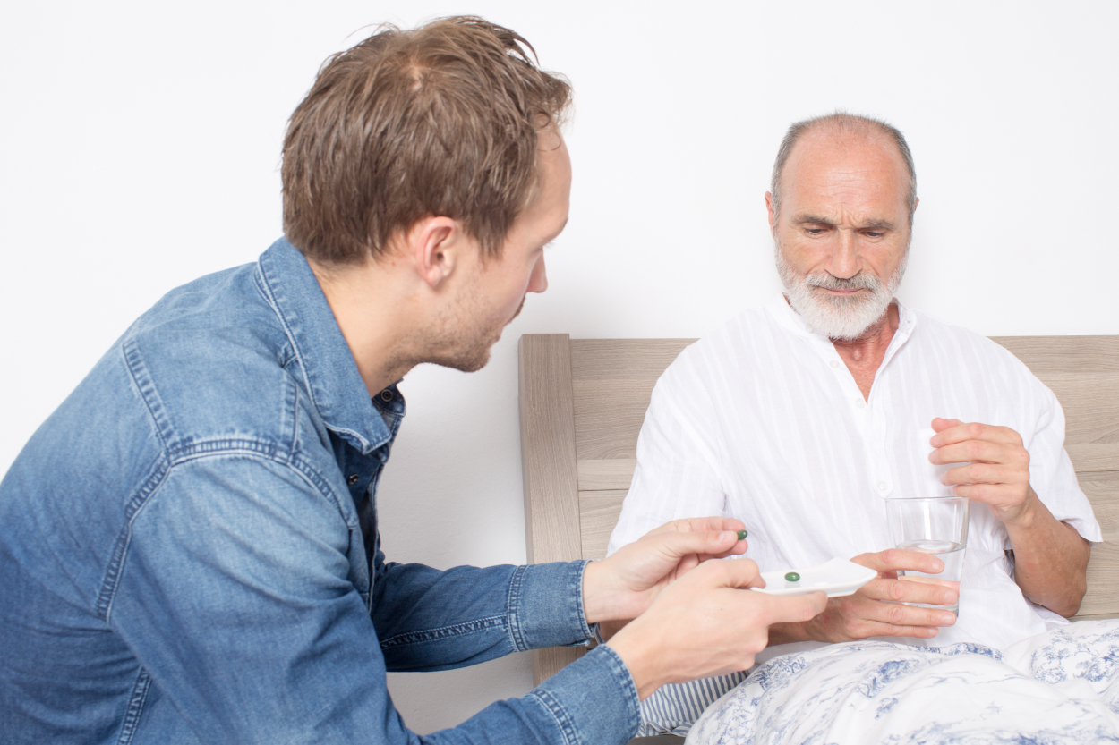 An older man in bed holding a glass of water; another man sits beside him holding a white plate with two pills