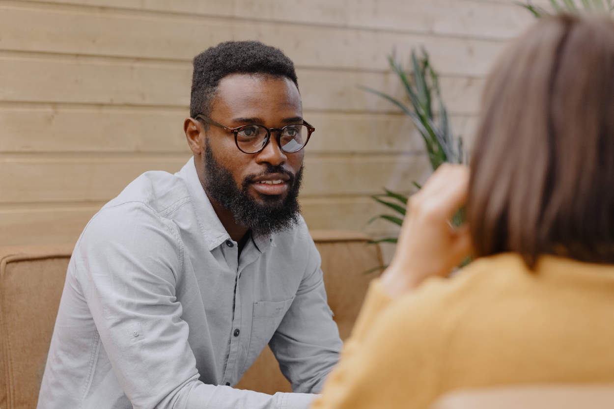 A black man wearing glasses sits across from another person. We only see the back of the other person who has a yellow shirt and shoulder-length brown hair. The man is sitting on a couch.